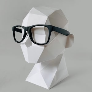 May include: Black-framed spectacles sit on a white, geometric paper sculpture of a head. The spectacles have a classic, rectangular shape. The sculpture is displayed against a plain, light grey background.