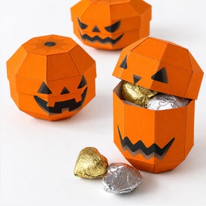Pode incluir: Três caixas de papel laranja em forma de abóbora com rostos de jack-o'-lantern pretos. Duas caixas estão fechadas e uma aberta, revelando doces embrulhados em papel dourado e prateado. Dois doces adicionais estão na superfície. Texto: DIGITAL DOWNLOAD, PDF & SVG FILES, NO PHYSICAL ITEM.