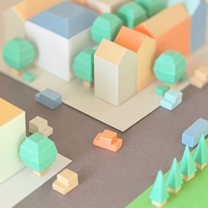 May include: A miniature paper craft city model featuring geometric buildings in pastel shades of peach, blue, and white. The scene includes small paper trees in mint green, tiny cars in orange and blue, and a grey road surface on a flat base.