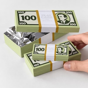 Puede incluir: Una caja rectangular y pilas más pequeñas diseñadas para parecer fajos de billetes de cien dólares. El papel verde y blanco presenta el texto PLAY CASH, el número 100 y un retrato. El interior de la caja está forrado con papel de aluminio.