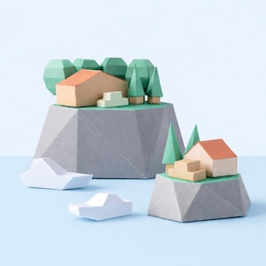 May include: A paper craft diorama featuring two gray geometric mountain bases, tan houses with orange roofs, green paper trees, and small white paper boats. The scene is constructed from folded cardstock, displaying sharp angles and clean lines.