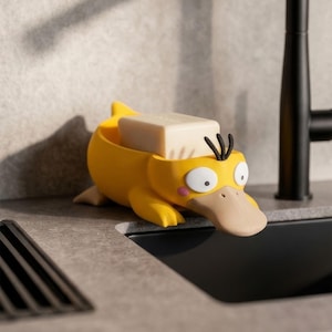 May include: A yellow soap dish shaped like a duck-billed creature with a wide tan beak, large white eyes, and three black hair tufts on its head. The dish holds a rectangular bar of off-white soap and features a textured, matte finish.