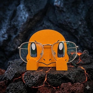 Könnte beinhalten: Ein leuchtend orangefarbener, abgerundeter Brillenhalter in Charakterform mit großen, stilisierten Augen und kleinen Händen am unteren Rand. Der Halter ist so gestaltet, dass er eine dünne, silberfarbene Metallbrille auf der Vorderseite trägt.