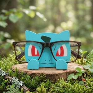 Puede incluir: Un soporte para gafas de color azul claro con forma de criatura, que presenta una marca verde oscuro en la frente y ojos rojos. El soporte está diseñado con dos pequeñas patas delanteras que sostienen unas gafas de montura marrón oscuro.