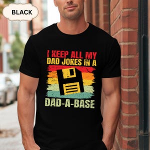 Puede incluir: Una camiseta negra de manga corta con un estampado gráfico que incluye el texto I KEEP ALL MY DAD JOKES IN A DAD-A-BASE. El diseño presenta un icono de disquete negro centrado sobre franjas horizontales en colores rojo, naranja, amarillo y verde azulado.