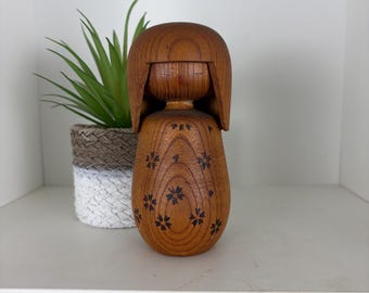 May include: A wooden Kokeshi doll with a rounded body and a simple, stylized face. The doll is made of light brown wood and has a dark floral pattern. The doll is displayed on a white shelf with a small potted plant.