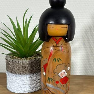 Könnte beinhalten: Eine traditionelle japanische Holzpuppe, bekannt als Kokeshi, mit einem schwarzen Haarknoten und einem bemalten Blumenkimono. Die Puppe steht auf einer Holzoberfläche.