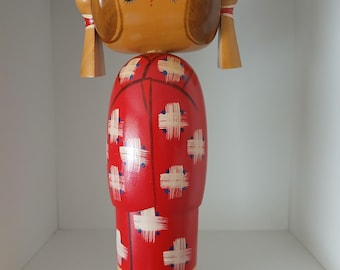 May include: A wooden Kokeshi doll with a rounded head and cylindrical body. The doll's head is light brown with painted facial features and hair. The red body is decorated with white cross-like patterns and blue dots. The doll is approximately 10 inches tall.
