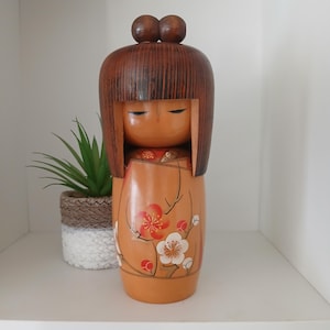 Könnte beinhalten: Kokeshi-Puppe aus Holz mit braunem Kopf und Haaren sowie einem beigefarbenen Körper, der mit roten und weißen Blumenmustern verziert ist. Die Puppe hat ein schlichtes, minimalistisches Design und steht auf einem weißen Regal.