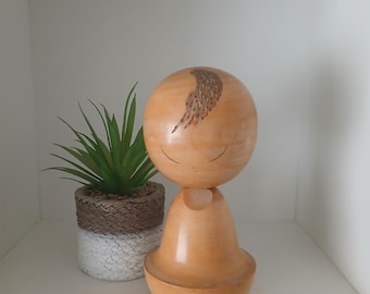 May include: A wooden doll with a round head and a pear-shaped body. The head has carved details and closed eyes. A small potted plant with green leaves sits next to the doll. The doll is displayed on a white shelf.