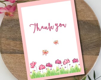 May include: A pink-bordered greeting card with the words "Thank you" in pink script. The card features watercolor illustrations of pink flowers, green grass, and butterflies. The card is on a wooden plate.
