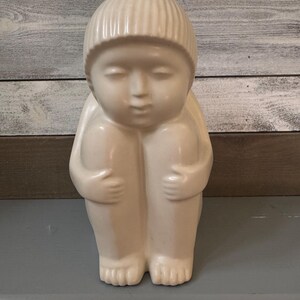May include: A white ceramic sculpture of a person sitting with their legs crossed and their arms wrapped around their legs. The person is wearing a hat.