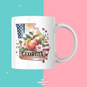 May include: White ceramic mug featuring a watercolor design of the state of Georgia. The design includes a peach, flowers, a bee, and the word "GEORGIA" on a wooden sign. The state outline incorporates the American flag.