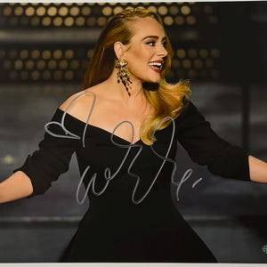 Op de afbeelding: Een gesigneerde foto van een vrouw met blond haar, gekleed in een zwarte jurk en gouden oorbellen. De foto heeft een handtekening met zilveren inkt over de voorkant. De vrouw lacht en kijkt opzij.