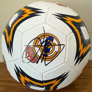Puede incluir: Un balón de fútbol blanco con un patrón repetitivo de gráficos en forma de rayo de color azul oscuro y amarillo. El centro del balón muestra un emblema circular con una corona roja y una firma en tinta negra sobre el diseño del logo.