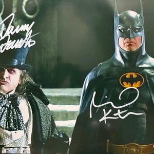 May include: A photograph featuring two characters from the Batman film series. The Penguin, in a top hat and cape, stands beside Batman in his iconic black suit with a yellow bat symbol. Both figures have white signatures.