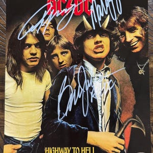 Puede incluir: Portada del álbum "Highway to Hell" de AC/DC con los miembros de la banda. La imagen está firmada con tinta plateada. La portada del álbum tiene un fondo oscuro con el logo de la banda en la parte superior. El texto "HIGHWAY TO HELL" está en la parte inferior.