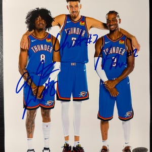 Puede incluir: Fotograf&iacute;a de tres jugadores de baloncesto con camisetas azules y naranjas de los Thunder. Cada camiseta tiene un n&uacute;mero y la palabra "Thunder". Los jugadores est&aacute;n de pie juntos, con un jugador abrazando a los otros dos. La foto est&aacute; firmada con tinta azul.