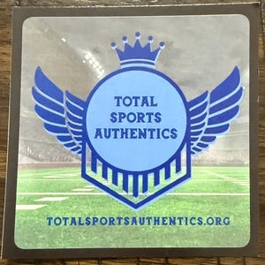 Puede incluir: Una pegatina cuadrada con el texto "TOTAL SPORTS AUTHENTICS" en un c&iacute;rculo azul, enmarcado por alas azules y una corona. El fondo muestra un campo de f&uacute;tbol. La direcci&oacute;n del sitio web est&aacute; en la parte inferior.