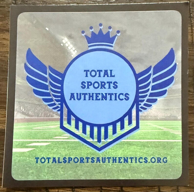 Puede incluir: Una pegatina cuadrada con el texto "TOTAL SPORTS AUTHENTICS" en un c&iacute;rculo azul, enmarcado por alas azules y una corona. El fondo muestra un campo de f&uacute;tbol. La direcci&oacute;n del sitio web est&aacute; en la parte inferior.