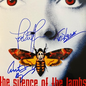 Puede incluir: Un cartel de la película "El silencio de los corderos" con un diseño de polilla con calavera. El título es rojo, con firmas en tinta azul. El fondo muestra un primer plano de un rostro con ojos rojos.