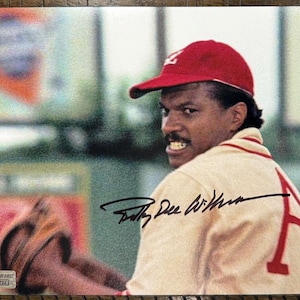 May include: A photograph of a man wearing a red baseball cap and a beige jacket with a red "A" on the back. The man is looking over his shoulder. The jacket has red trim. The image is signed with a black pen.