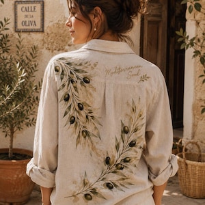 Peut inclure: Une chemise beige à manches longues boutonnée avec un motif peint de branches d'olivier et d'olives vert foncé au dos. Le texte "Mediterranean Soul" est imprimé en lettres dorées près de l'épaule, accompagné d'une petite icône de feuille.