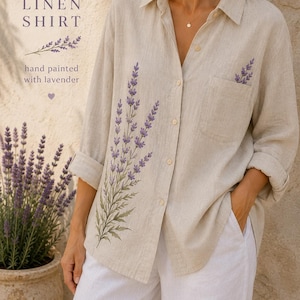 Puede incluir: Una camisa de lino beige con ramas de lavanda pintadas a mano en la parte delantera y en el bolsillo. La camisa tiene cuello, cierre de botones y mangas largas. El texto dice: LINEN SHIRT, hand painted with lavender.