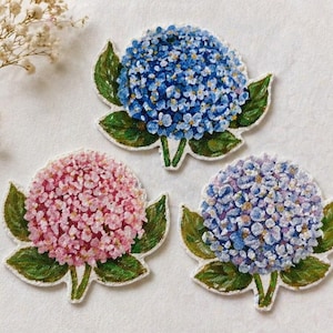 Puede incluir: Tres parches bordados con flores de hortensia en tonos azules, rosas y morados. Cada parche presenta un diseño floral detallado con hojas y tallos verdes, sobre un fondo blanco. Los parches son probablemente para fines decorativos.