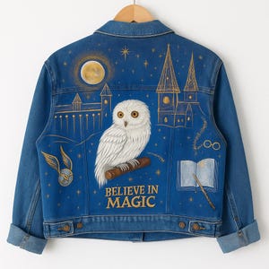 Puede incluir: Chaqueta vaquera azul con un diseño pintado a mano que presenta un búho blanco, una luna llena y un castillo. La frase "BELIEVE IN MAGIC" está escrita en letras doradas. La chaqueta tiene botones y mangas dobladas.