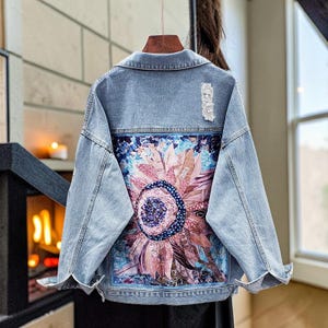 Könnte beinhalten: Hellblaue Jeansjacke mit einem großen, farbenfrohen Sonnenblumenmuster auf dem Rücken. Die Sonnenblume hat rosa, blaue und violette Farbtöne. Die Jacke hat ein Distressed-Detail und einen klassischen Kragen.