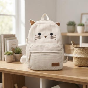 Può includere: Uno zaino in velluto a coste color crema con un design ispirato a un gatto, dotato di due orecchie a punta sulla parte superiore, due occhi neri circolari e tre baffi ricamati su ogni lato. Include una tasca frontale con zip e un'etichetta marrone.