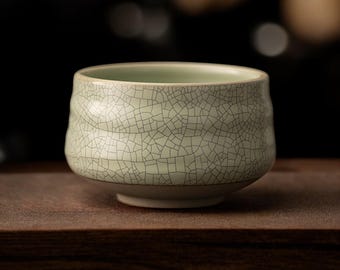 May include: A pale mint green ceramic tea bowl featuring a distinct dark crackle glaze pattern across its exterior surface. The bowl has a rounded, slightly undulating shape with a smooth rim and a solid, narrow base, showcasing a textured finish.