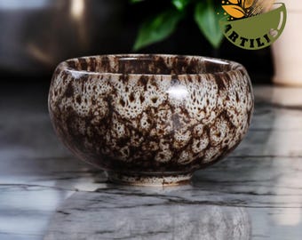 May include: A ceramic bowl featuring a mottled brown and white glaze with a distinct speckled pattern. The bowl has a rounded shape with a small footed base. A logo with the text ARTILIS and a bamboo whisk graphic is visible in the upper right corner.