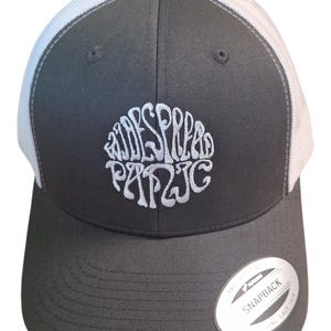 May include: A dark gray trucker hat with a white mesh back and a curved brim. The front of the hat features white embroidered text in a circular design. A small, white tag with black text is on the side.