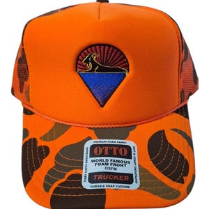 May include: An orange camouflage trucker hat with a foam front and a triangular embroidered patch featuring a black goat silhouette against a red sun and blue horizon. A sticker on the brim reads OTTO WORLD FAMOUS FOAM FRONT OSFM TRUCKER.