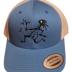 May include: A blue baseball cap with a tan mesh back and an embroidered black figure playing a banjo. The front features a circular sticker that reads THE AUTHENTIC YUPOONG SNAPBACK YUPOONG SINCE 1974. The cap has a structured crown and a flat brim.