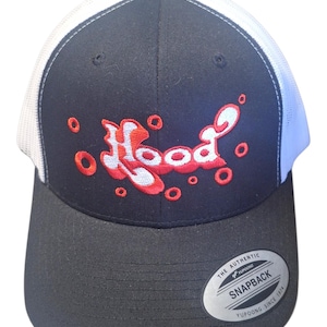 Könnte beinhalten: Eine schwarz-weiße Trucker-Cap mit schwarzer Vorderseite, auf der das Wort Hood in Weiß und Rot gestickt ist, umgeben von kleinen roten Kreisen. Die Kappe hat einen schwarzen Schirm und einen silbernen Aufkleber mit der Aufschrift The Authentic Yupoong Snapback.