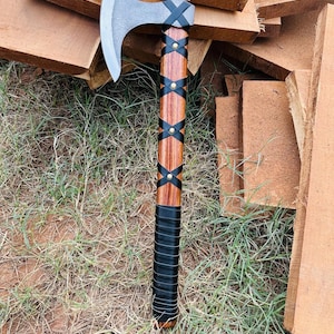Könnte beinhalten: Eine handgefertigte Axt mit einer silbernen Klinge und einem Holzgriff. Der Griff ist mit schwarzem Leder umwickelt und mit schwarzen Riemen und Messingnieten befestigt. Die Axt ist zum Hacken und Schneiden bestimmt.