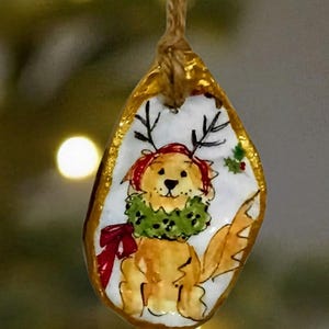 以下が含まれることがあります： 手描きのクリスマスオーナメント。赤い帽子と角、緑のリースをつけたゴールデンレトリバーが描かれています。オーナメントは金色の縁取りで、天然の麻ひもループで吊り下げられています。お祝いのホリデーデコレーション。