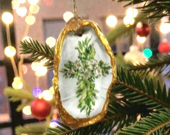 Puede incluir: Adorno navideño pintado a mano con forma de concha de ostra. El adorno presenta un borde dorado y un interior blanco con un diseño de cruz floral verde y blanco. Cuelga de un árbol de Navidad con adornos rojos y verdes.