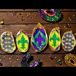 Könnte beinhalten: Handbemalte Austernschalen mit goldenen Rändern, die Mardi Gras-Designs zeigen. Die Schalen zeigen Fleur-de-Lis- und Rautenmuster in Lila, Grün und Gelb. Eine Glitzermaske und dekorative Akzente vervollständigen das festliche Thema.