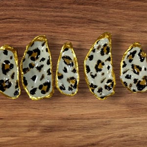 May include: Five oyster shell dishes with a white and black leopard print pattern and gold trim. The shells are arranged in a row on a wooden surface. The shells vary in size.
