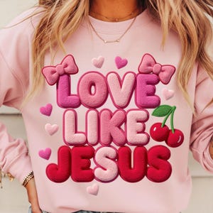 Può includere: Felpa rosa chiaro con la scritta "LOVE LIKE JESUS" in un font a bolle 3D. Il design include fiocchi, cuori e ciliegie rosa. La felpa è di colore rosa chiaro.
