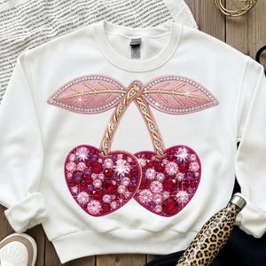 Può includere: Felpa bianca con un grande design di ciliegie decorate. Le ciliegie sono a forma di cuore e riempite di strass rossi e rosa. I gambi e le foglie sono dettagliati con ricami dorati e rosa, creando un design scintillante.