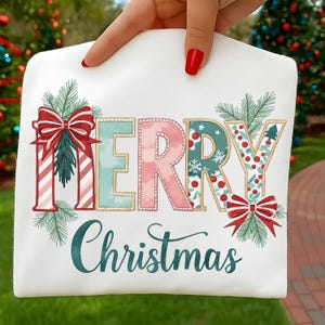 Może przedstawiać: Biała koszulka z napisem "MERRY Christmas" w świątecznym wzornictwie. Litery zdobione są kokardkami, paskami i świątecznymi wzorami. Zielone gałązki sosny i turkusowa czcionka skryptowa dopełniają projekt.