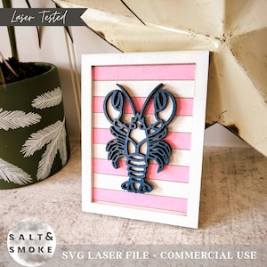 May include: A wooden framed artwork with a navy blue lobster design. The lobster is set against a pink and white striped background within a white frame. The words "Laser Tested" are at the top.