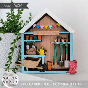 May include: A wooden potting shed ornament with a blue frame, brown walls, and a white roof. The shed features miniature gardening tools, pots, and a wheelbarrow. The text "THE POTTING SHED" is above the shelves. Red polka dot wellington boots are on the right.