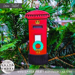 Può includere: Un ornamento natalizio in legno rosso a forma di cassetta postale britannica, con una corona d'oro e una ghirlanda verde. L'ornamento è appeso a un albero di Natale. Il testo "Laser Tested" è visibile.