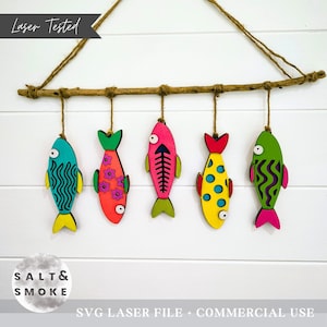 May include: A decorative wall hanging featuring five colorful wooden fish suspended from a natural branch. Each fish is painted in vibrant hues with unique patterns, including stripes, flowers, and polka dots. The text "Laser Tested" is visible.